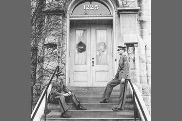 Two men in military garb talking to eachother in front of a door with the street number 1225. Links to Gifts by Will